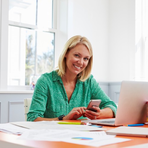Woman Sitting At Desk Using Mobile Phone In Home Office